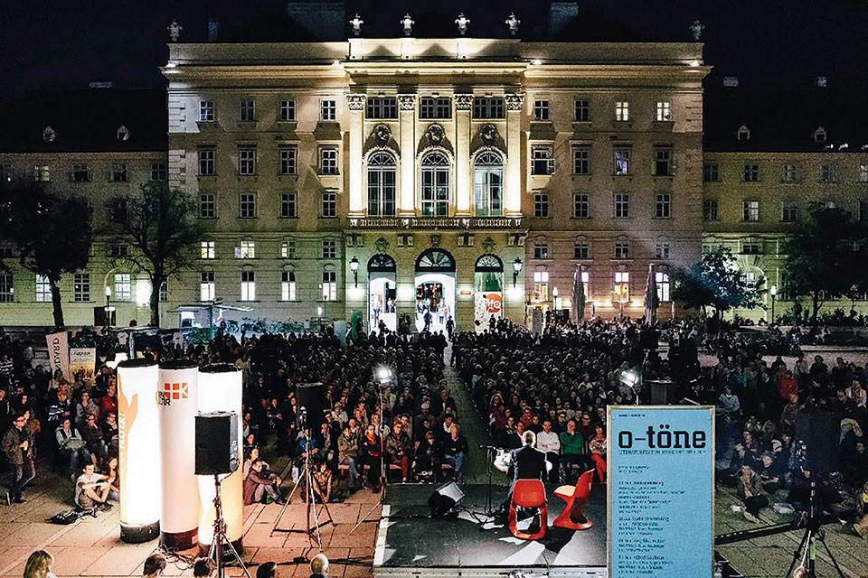 Looking from behind an outdoor stage towards a large crowd assembled to watch a performance with a large building in the background