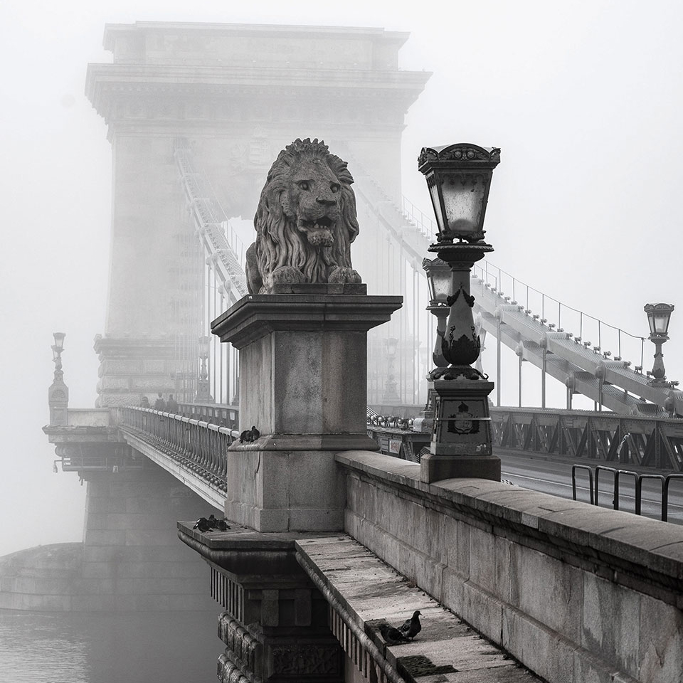 A stone lion adorns the entrance to a bridge that stretches into the background which is swaddled in fog