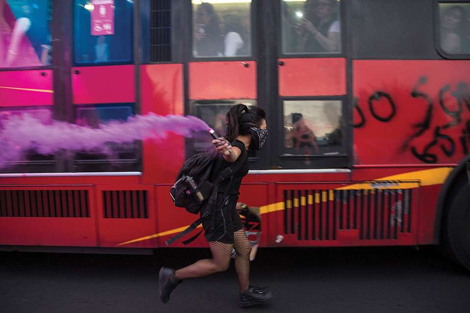 A woman masked in a black bandana runs alongside a bus covered with graffiti carrying a purple flare