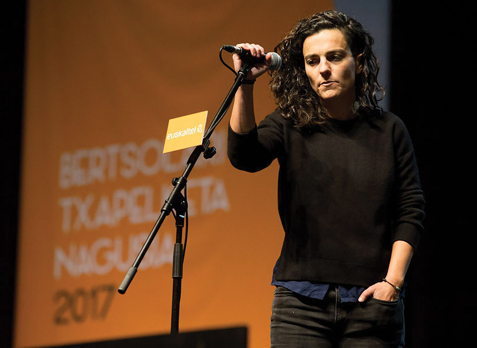 A woman dressed all in black holds a microphone in a stand on a stage