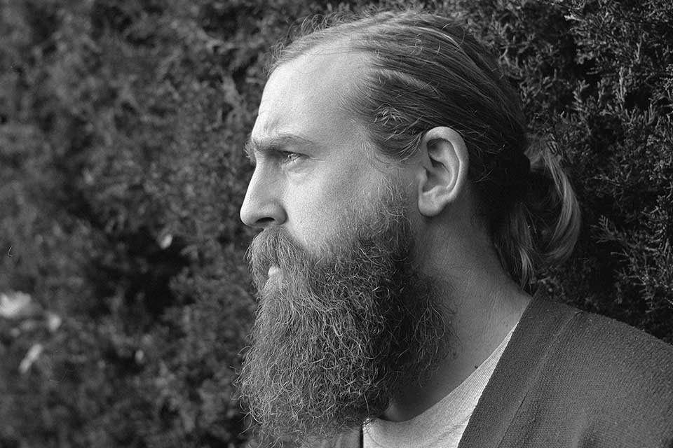 A black and white photograph of a bearded man standing in front of a tall hedge