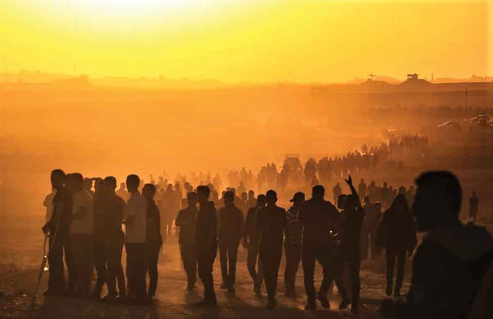 A photograph of a throng of people walking on a dusty landscape at sunset