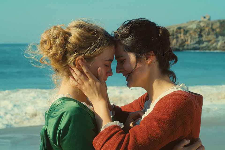 A still photograph from Portrait of a Lady on Fire that features two women standing on a beach, holding one another and pressing foreheads