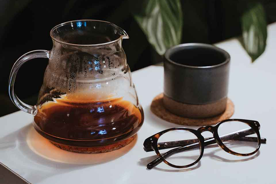 A photograph of a clear tea pot, a tea mug, and a pair of glasses on a table