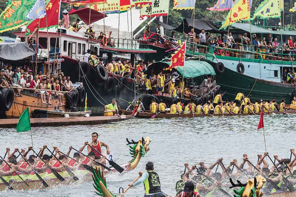 A photograph of a raucous public festival where teams of rowers in dragon boats traverse a river