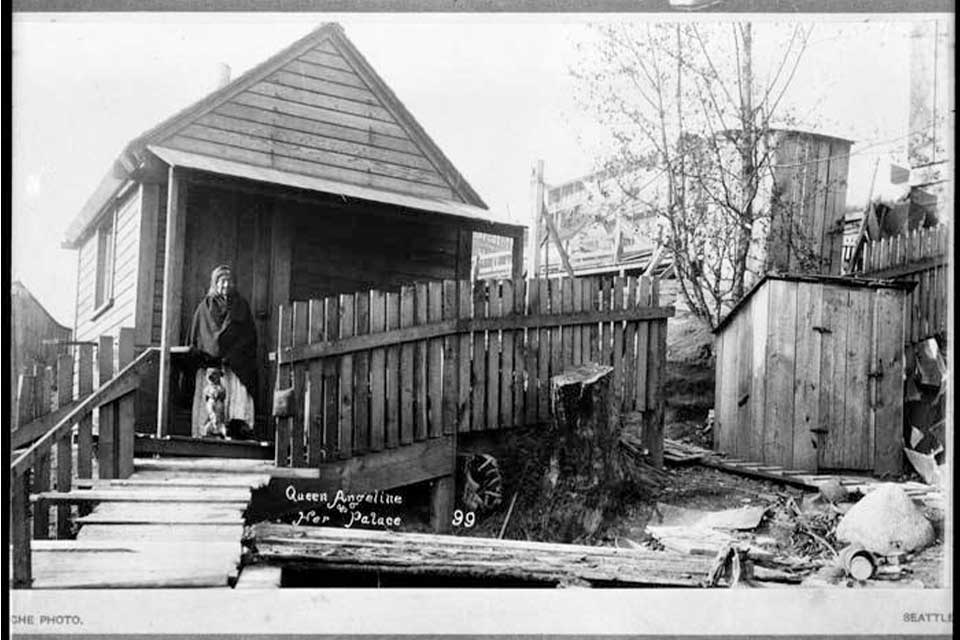 An historical black and white photograph of an indigenous woman standing on the porch of a simple home newly constructed from lumber