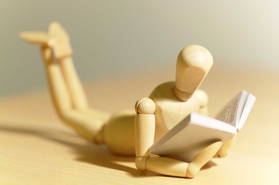 A close-up photograph of a small, wooden figure reading a tiny book.