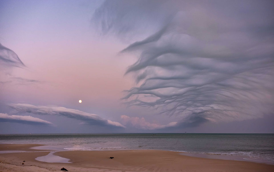 A beach dissolves into a shallow sea, beneath a purpled sky laced with wispy clouds