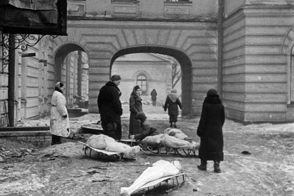 A black and white photograph of a number of people, all in heavy coats, standing in an alley way with a number of bodies wrapped in shrouds