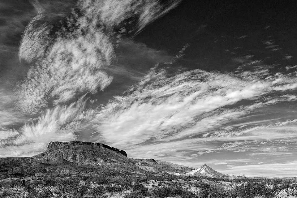 A black and white photograph of a cloud streaked sky above a mesa and the scrublands below