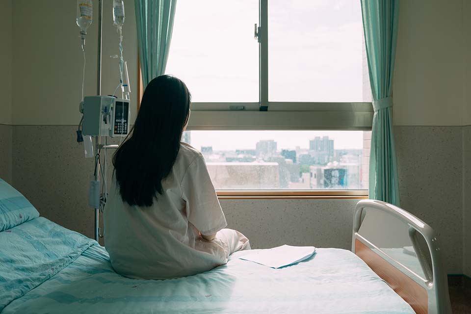 A photograph of a woman in a hospital gown seated away from the camera, looking out a nearby window