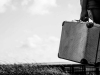 A black and white photograph of a hand attached to a figure in a business suit, holding a weathered suitcase