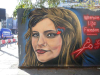 A mural of a woman's face with a pair of scissors near her head rendered in dark red. Text reads: Woman. Life. Freedom. 