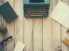 A typewriter, notepad, books, and glasses circle an empty spot on a rough woodgrain table