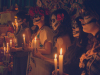 A group of people with their faces painted white like skulls hold candles before an altar