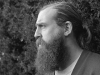 A black and white photograph of a bearded man standing in front of a tall hedge