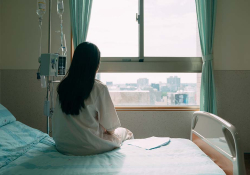 A photograph of a woman in a hospital gown seated away from the camera, looking out a nearby window