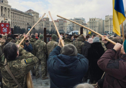 A photograph of a group of three people holding long wooden poles to their face in a crowd of protesters