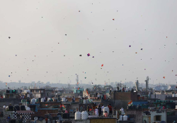 A distant shot of a cityscape. Kites dot the sky are rising into the air.