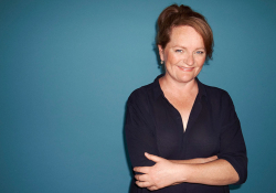 A photograph of Dorthe Nors, who smiles at the viewer, arms folded, against a dark blue background