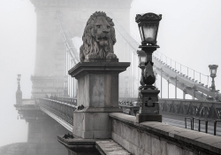 A stone lion adorns the entrance to a bridge that stretches into the background which is swaddled in fog