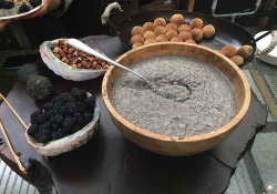 A spoon, partially submerged in a grain, rests on the edge of a wooden bowl, which itself is on a table with fruits and nuts